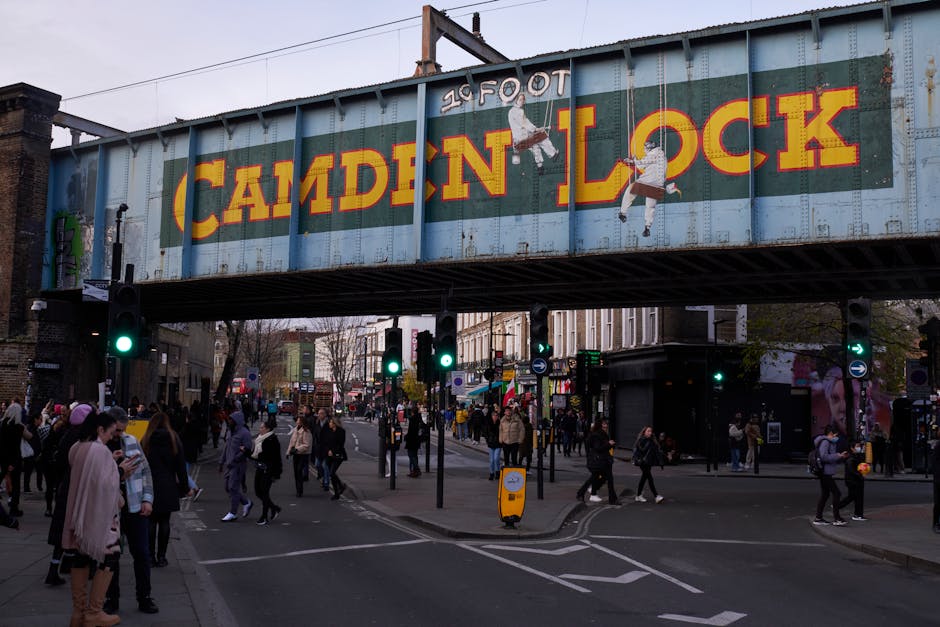 A large, weathered railway bridge with a faded greenish-blue paint, featuring the bold, yellow lettering 'Camden Lock' on its side in a historic district of Camden, London. The bridge spans over a bus