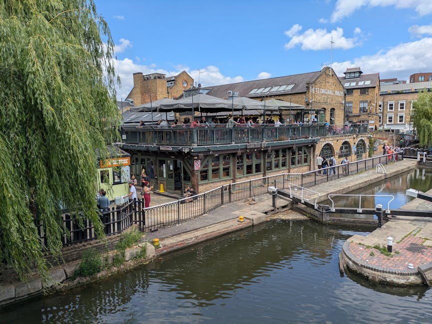 The image depicts a multi-level brick building situated alongside a canal, with a large outdoor terrace on the upper level covered by a dark canopy and filled with patio furniture and people dining or socialising. The building has large arched windows on the ground floor, and the canal is separated from the building by a metal railing and a paved walkway where a few individuals are walking or standing. A small green vintage-style van is parked near the building, with some boxes and packing materials nearby, possibly indicating a loading or unloading area for a house relocation service. The canal water reflects the blue sky and surrounding structures, and a narrow bridge with railings crosses a section of the waterway, suggesting access for pedestrians. Trees with lush green foliage partly frame the scene, and the background features a mix of older and modern brick buildings under a partly cloudy sky. Camden Man and Van's services may involve logistics around packing, furniture transport, and home moves in this area, as implied by the context of house removals and relocation within Camden.