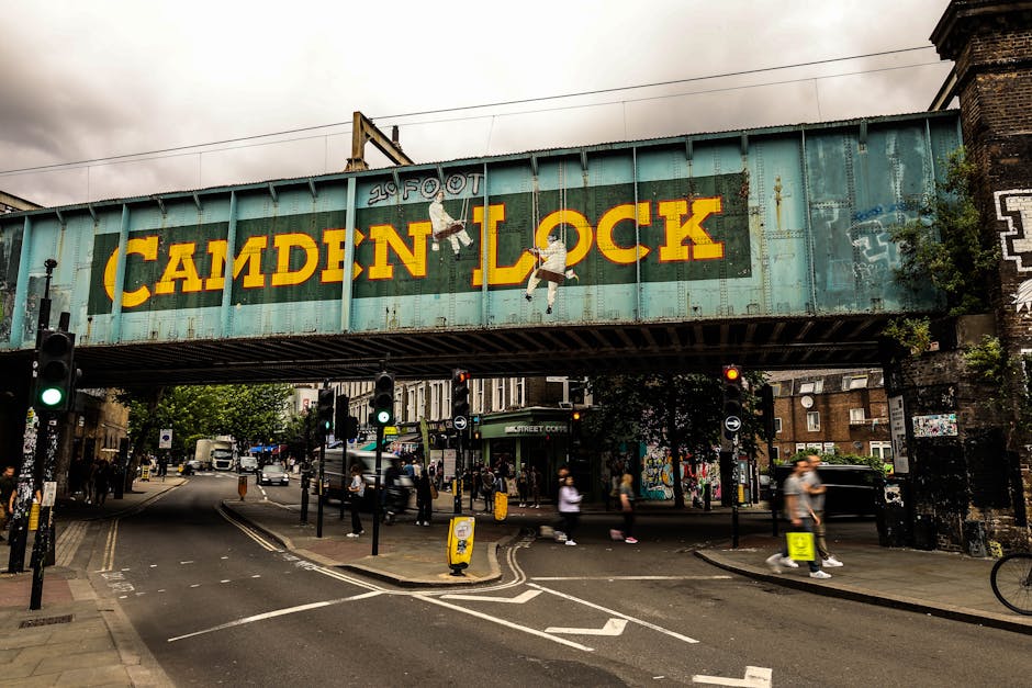 A large, weathered blue metal bridge structure spans over a busy street in Camden, London, with the words 'CAMDEN LOCK' painted in bold yellow letters. The bridge features faded skateboarding illustrations and the mention of '20 FOOT' in smaller text. Below, traffic lights and pedestrian crossings control movement of vehicles and pedestrians, some of whom are blurred due to motion, indicating active foot traffic. The street is lined with small shops, and within the scene, there are various packages, cardboard boxes, and wrapped items, possibly being prepared for a home relocation involving furniture transport. Camden Man and Van's moving services are suggested by the context of furniture and packing materials in the vicinity of the street, which is part of the urban environment seen in the image. The overall scene highlights the logistical aspects of packing, loading, and moving within a vibrant city setting, with natural lightning illuminating the area under an overcast sky.