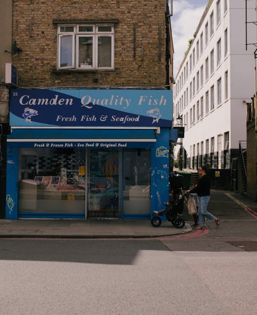 The image shows a street scene outside a fish and seafood shop called 'Camden Quality Fish', with a blue storefront sign and large display window. Above the shop, there is a brick residential building with a window on the upper floor. On the pavement in front of the shop, a woman is pushing a black stroller with a small child inside, while a man wearing a black shirt and light-colored pants walks beside her, carrying a brown paper shopping bag. The man appears to be in the process of loading or unloading items, potentially involving house or flat move logistics. Nearby, there are black wheelie bins and a narrow alleyway leading between buildings, suggesting an urban environment suitable for residential or commercial relocations. The scene is set during daylight, with natural light illuminating the street and storefront, capturing a typical day in Camden with opportunities for furniture transport, packing, or home relocation activities. Camden Man and Van is known for their removals services tailored to busy city neighborhoods like NW1 near Camden Market.