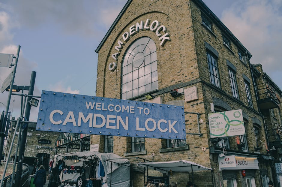 The exterior of a three-story brick building with large arched windows and black metal balconies, situated on a busy street near Camden Market NW1 in Camden. A large blue sign with white lettering reading 'Welcome to Camden Lock' is prominently displayed in front of the building. Below this, a smaller sign in a matching blue background states 'Camden Lock' with additional text about lock offices and shops to rent. The street scene includes pedestrians, market stalls with fabric and clothing items, and various signs and advertisements. Visible equipment such as a trolley and cushioning blankets are arranged nearby, suggesting a home relocation or furniture transport process is underway. The lighting appears natural, with an overcast sky overhead, emphasizing a professional moving environment connected to house removals and packing and loading activities facilitated by Camden Man and Van, typically operating in London’s Camden area.