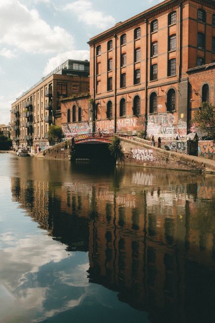 A view of a historic brick building adjacent to Camden's canal with a small stone bridge crossing over the water, which reflects the building and bridge. The building features multiple large arched windows and several floors, with some graffiti visible on its lower part. To the left, modern multi-storey structures with glass balconies line the street, indicating an urban setting suitable for house removals and furniture transport. The canal's calm water captures the detailed reflection of the building, bridge, and partly cloudy sky above. This scene illustrates an area where house removals and relocation services by Camden Man and Van often operate, highlighting the logistical environment and typical urban infrastructure involved in home relocation projects, including loading areas near the water's edge and potential access points for moving vehicles.