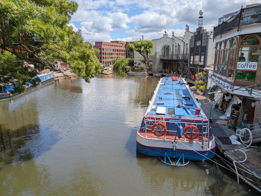 A narrow canal scene during daytime with a blue and white houseboat moored alongside a small walkway adjacent to commercial buildings, including a café with large glass windows and an outdoor seating area. The houseboat is secured to the quay with ropes and features safety equipment such as orange life rings on the stern. Nearby, a cluster of cardboard boxes, packing materials, and a small plastic container can be seen on the pavement, indicating packing or loading activities related to home relocation. The scene is illuminated by natural daylight with partly cloudy skies, and a large leafy tree extends over the canal, providing shade. Visible in the background are modern red-brick buildings and trees, emphasizing an urban riverside environment consistent with residential or commercial moving logistics facilitated by Camden Man and Van, whose services include furniture transport and packing during house removals along the Regents Canal area.
