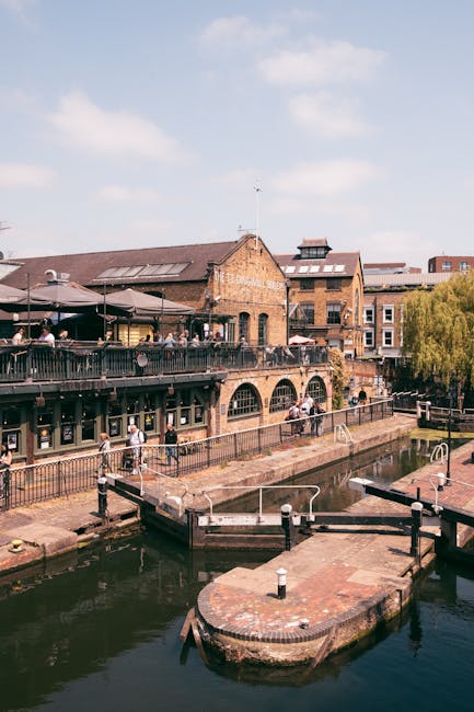 A view of a historic brick building adjacent to Camden's canal with a small stone bridge crossing over the water, which reflects the building and bridge. The building features multiple large arched windows and several floors, with some graffiti visible on its lower part. To the left, modern multi-storey structures with glass balconies line the street, indicating an urban setting suitable for house removals and furniture transport. The canal's calm water captures the detailed reflection of the building, bridge, and partly cloudy sky above. This scene illustrates an area where house removals and relocation services by Camden Man and Van often operate, highlighting the logistical environment and typical urban infrastructure involved in home relocation projects, including loading areas near the water's edge and potential access points for moving vehicles.